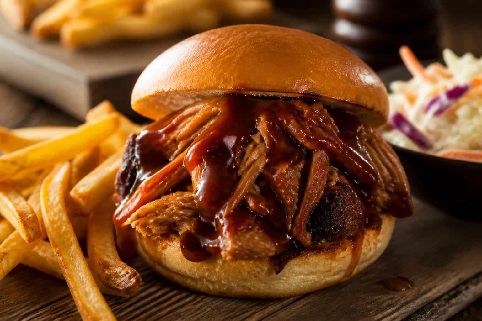 A BBQ pulled pork sandwich with barbecue sauce on a bun, served with French fries and coleslaw on a wooden table.