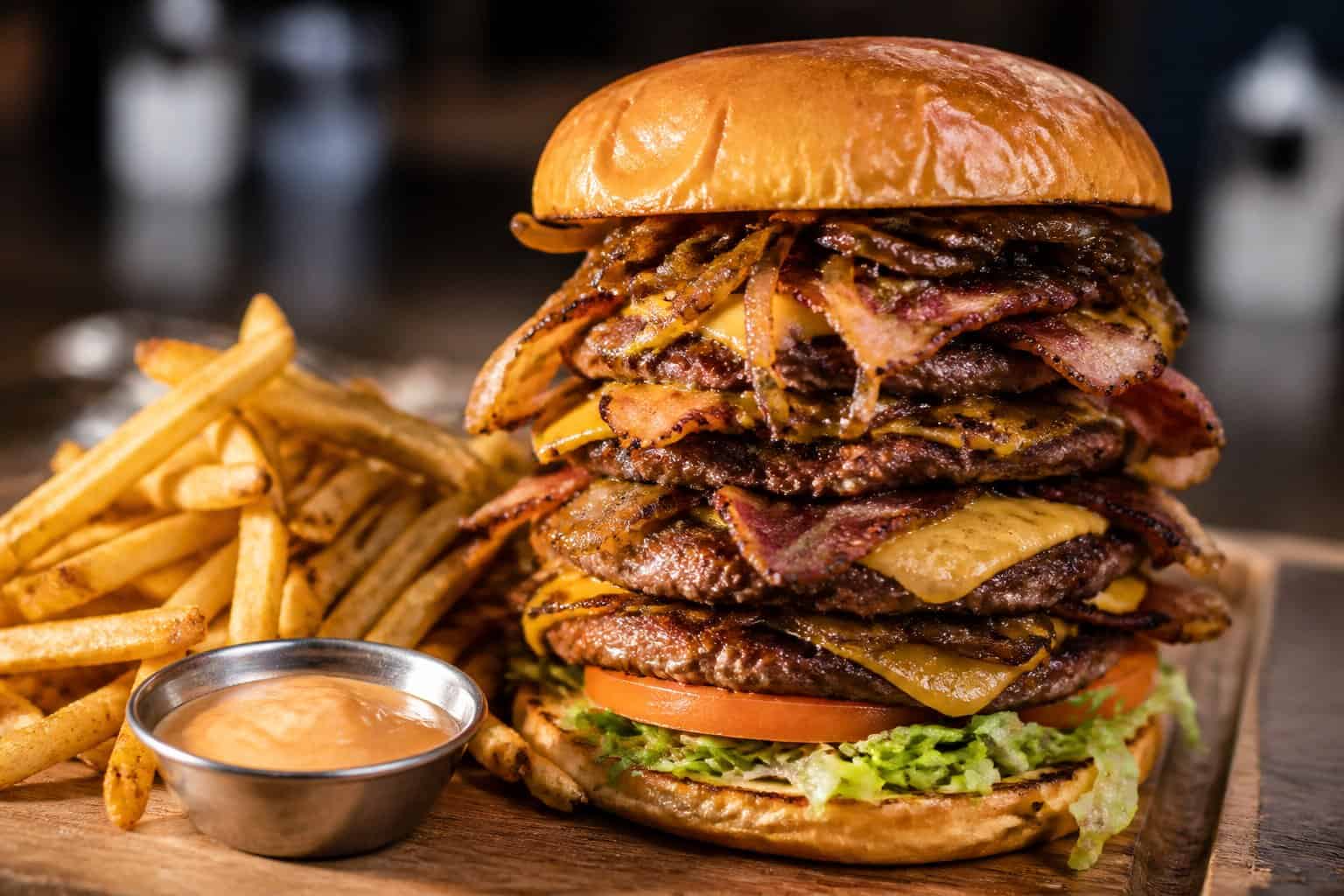A large, multi-layered burger with beef patties, cheese, bacon, lettuce, tomato, and onions on a brioche bun, served with French fries on a wooden board.