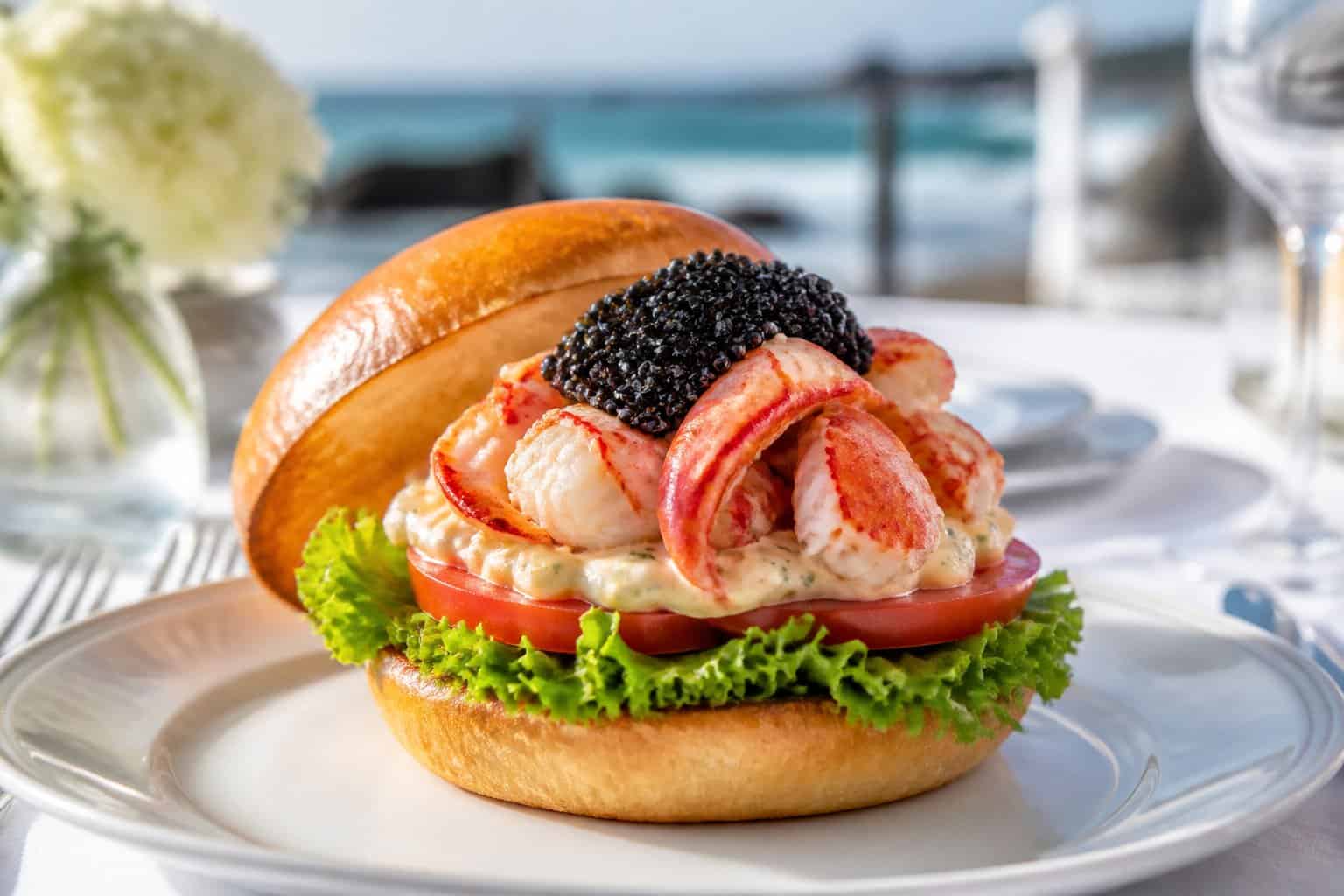 A lobster burger topped with black caviar on a toasted bun, served on a white plate with a blurred ocean background.