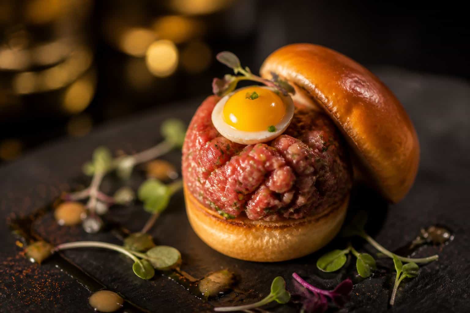 A close-up of a Wagyu tartare slider topped with a quail egg on a dark serving board with garnishes.