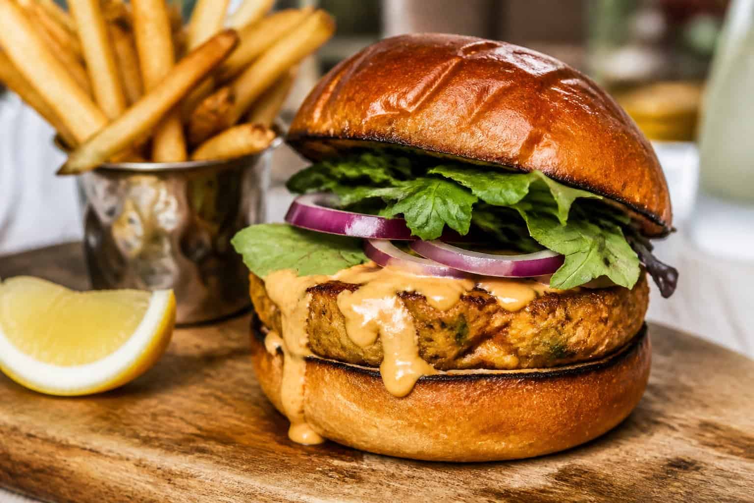 A smoked sablefish burger with citrus aioli on a toasted bun, served with fries and a lemon wedge on a wooden board.
