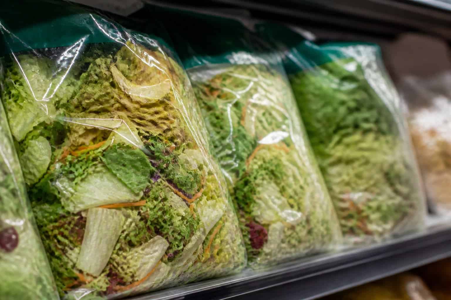 Close-up of several bagged salad mixes on a supermarket shelf showing wilted and browning lettuce leaves inside clear packaging.