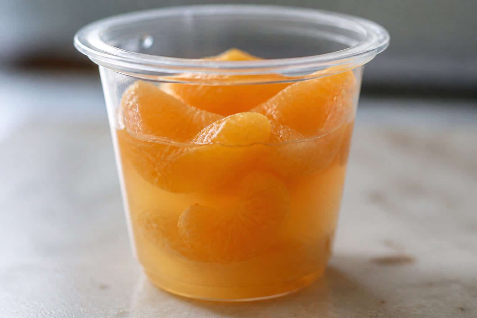 A clear plastic cup filled with mandarin orange segments in syrup on a neutral surface.