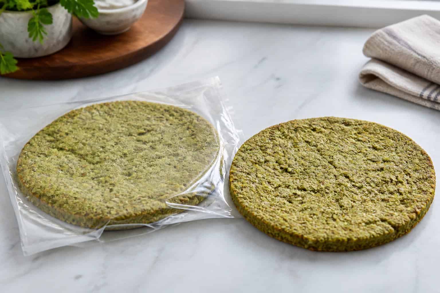 A green broccoli and kale pizza crust in plain packaging on a kitchen countertop.