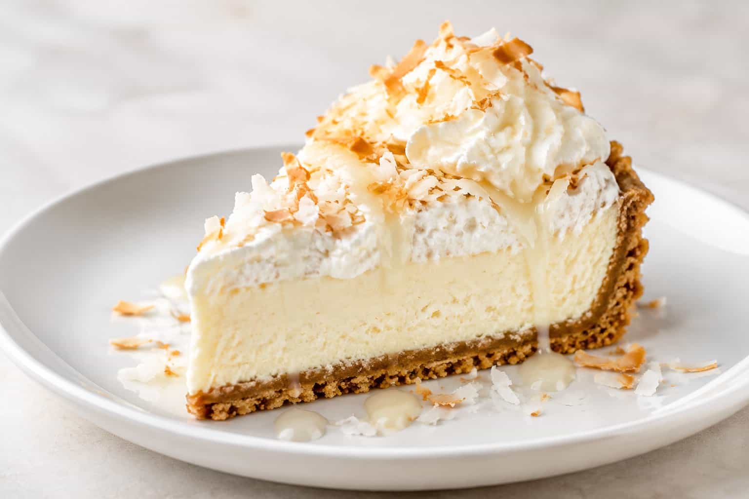 A slice of coconut cream pie cheesecake topped with whipped cream and toasted coconut on a white plate.