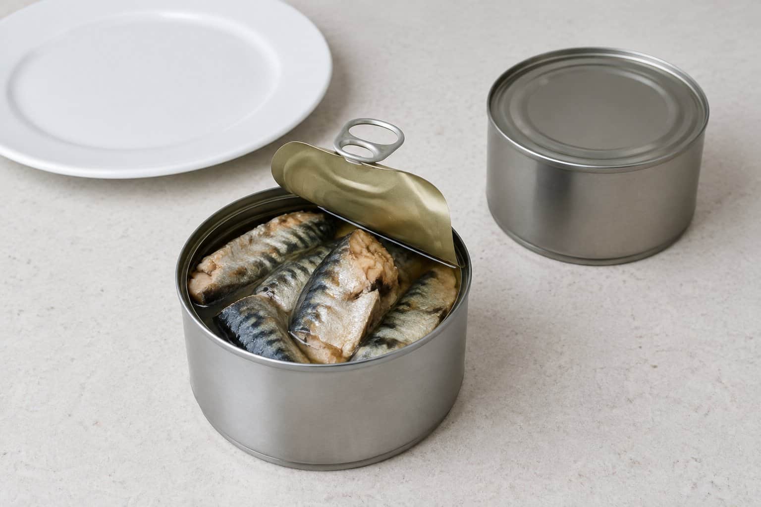 An opened can of mackerel beside a plate and an unopened can on a kitchen counter.