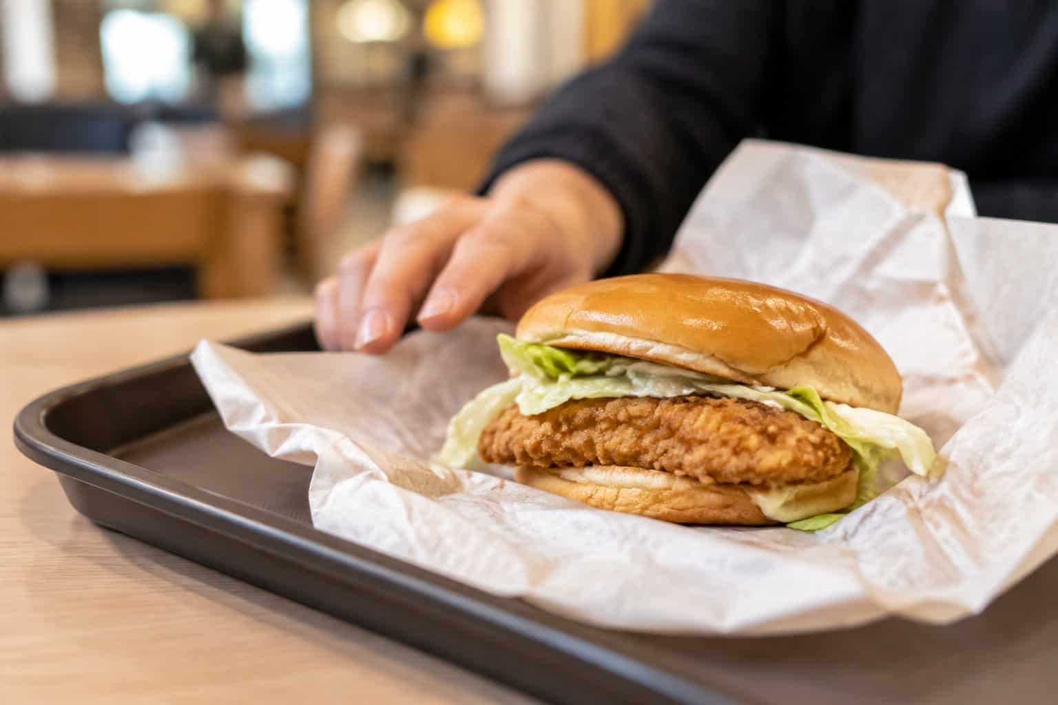 A fried chicken sandwich in an open wrapper on a tray as a customer pushes it aside.
