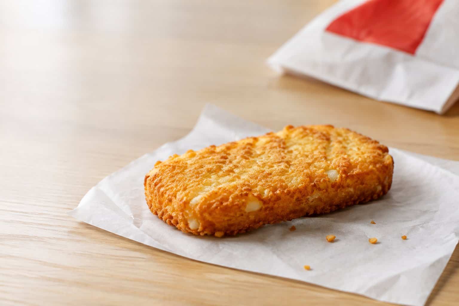 A golden-brown hash brown on a white wrapper beside a paper bag.
