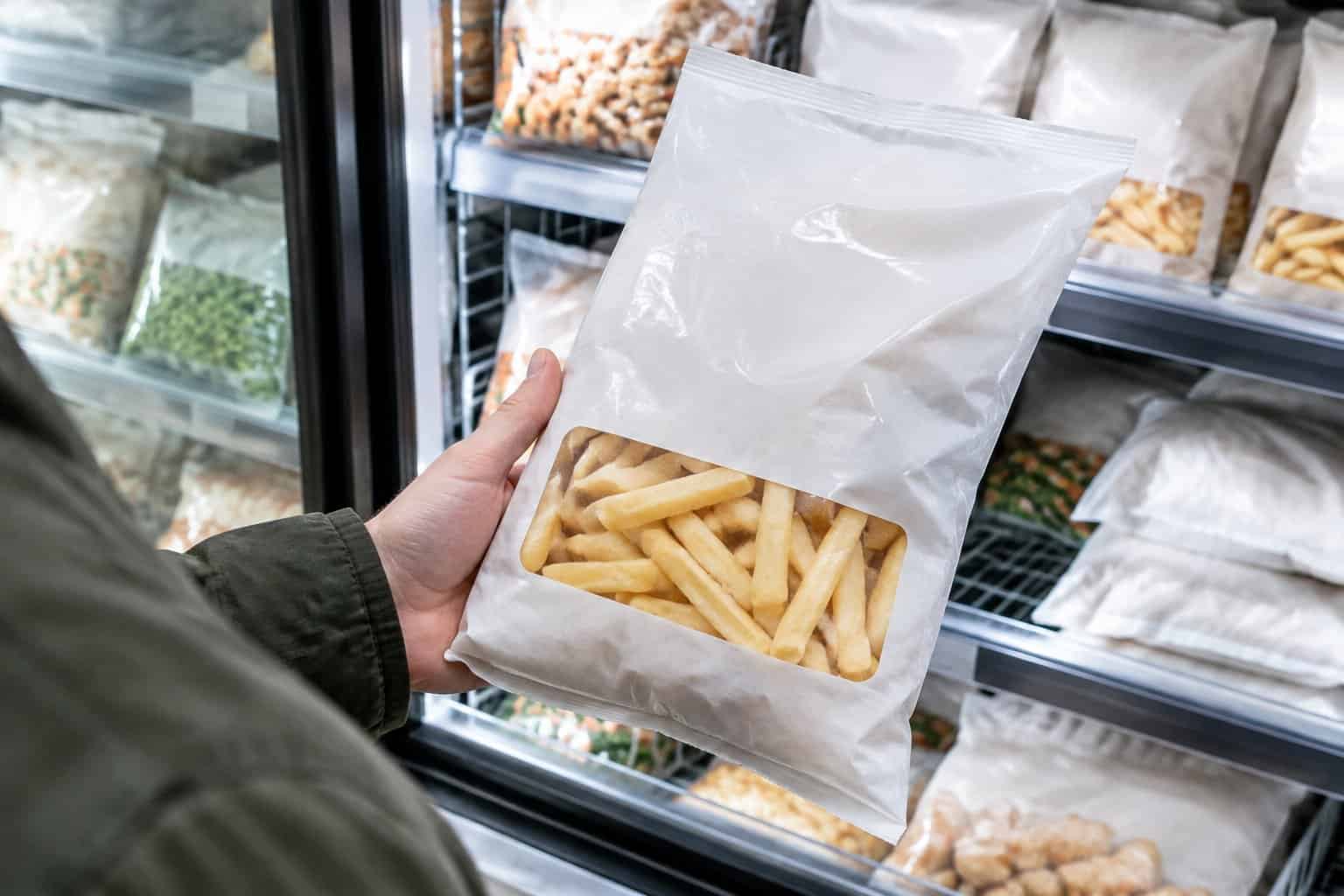 A shopper holds a bag of frozen French fries beside a supermarket freezer.