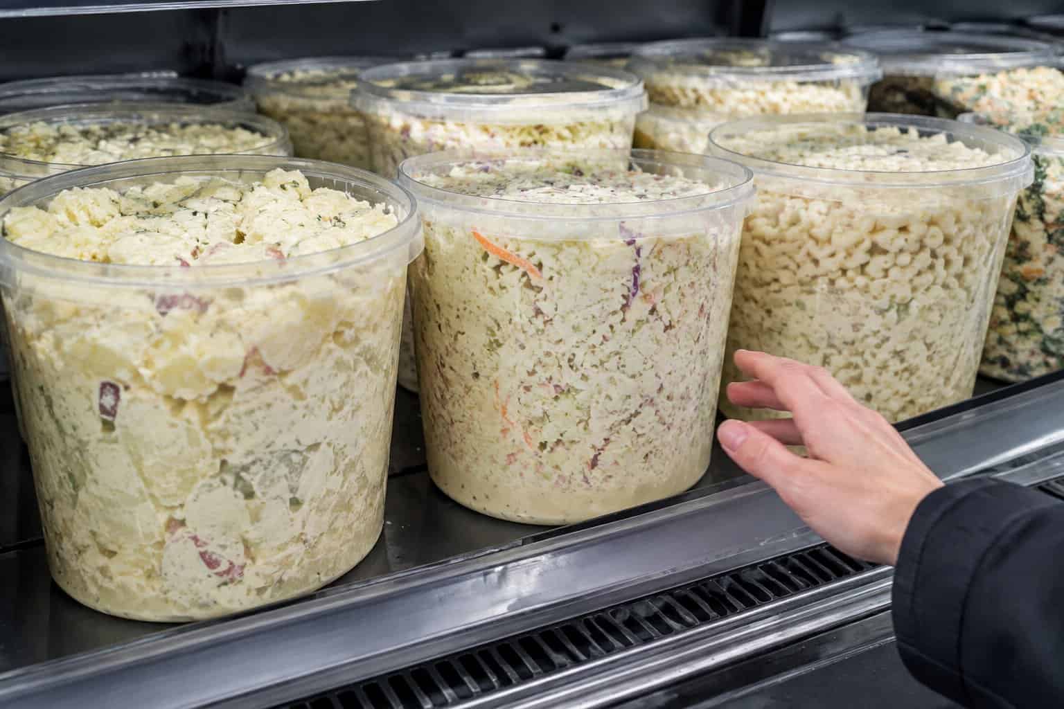 Large tubs of prepared salads in a supermarket refrigerated display as a shopper reaches toward them.