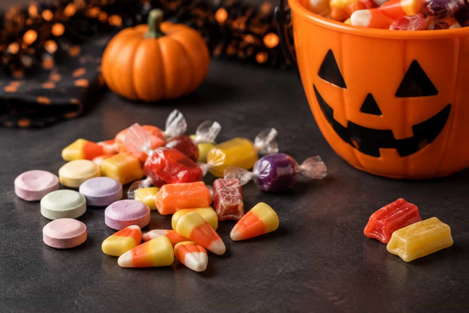 An assortment of Halloween candies sits beside a trick-or-treat bucket and a small pumpkin.
