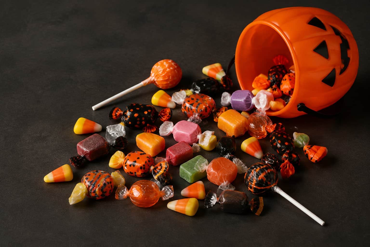 An orange pumpkin bucket spills assorted Halloween candies across a dark tabletop.